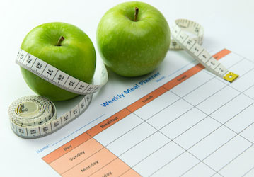 Apples and measuring tape sitting on paper with a weekly meal plan grid highlight healthy fueling for military fitness 