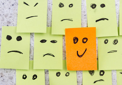 One orange post-it with happy face drawn on it in the middle of many yellow post-its with sad or mad faces on them