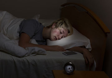 Teen boy sleeping in a dark room next to lit phone screen shows the importance of sleep for health  mental wellness  and perf