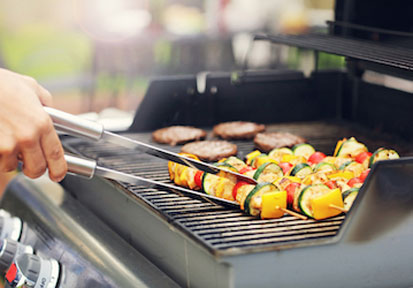 Person using tongs to turn vegetables and hamburgers on grill