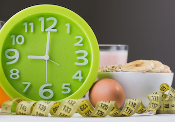 Green analog clock next to a tape measure  egg  and bowl of oatmeal indicates the benefit of nutrient timing for optimal perf